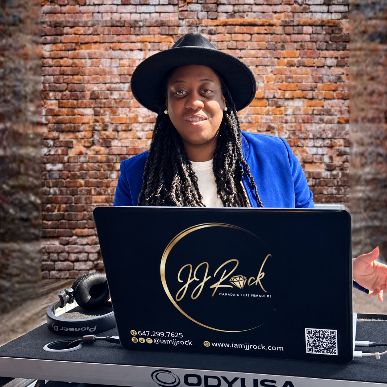 JJ Rock, founder of JJ Rock Entertainment Group, smiling behind her DJ setup in front of a brick wall